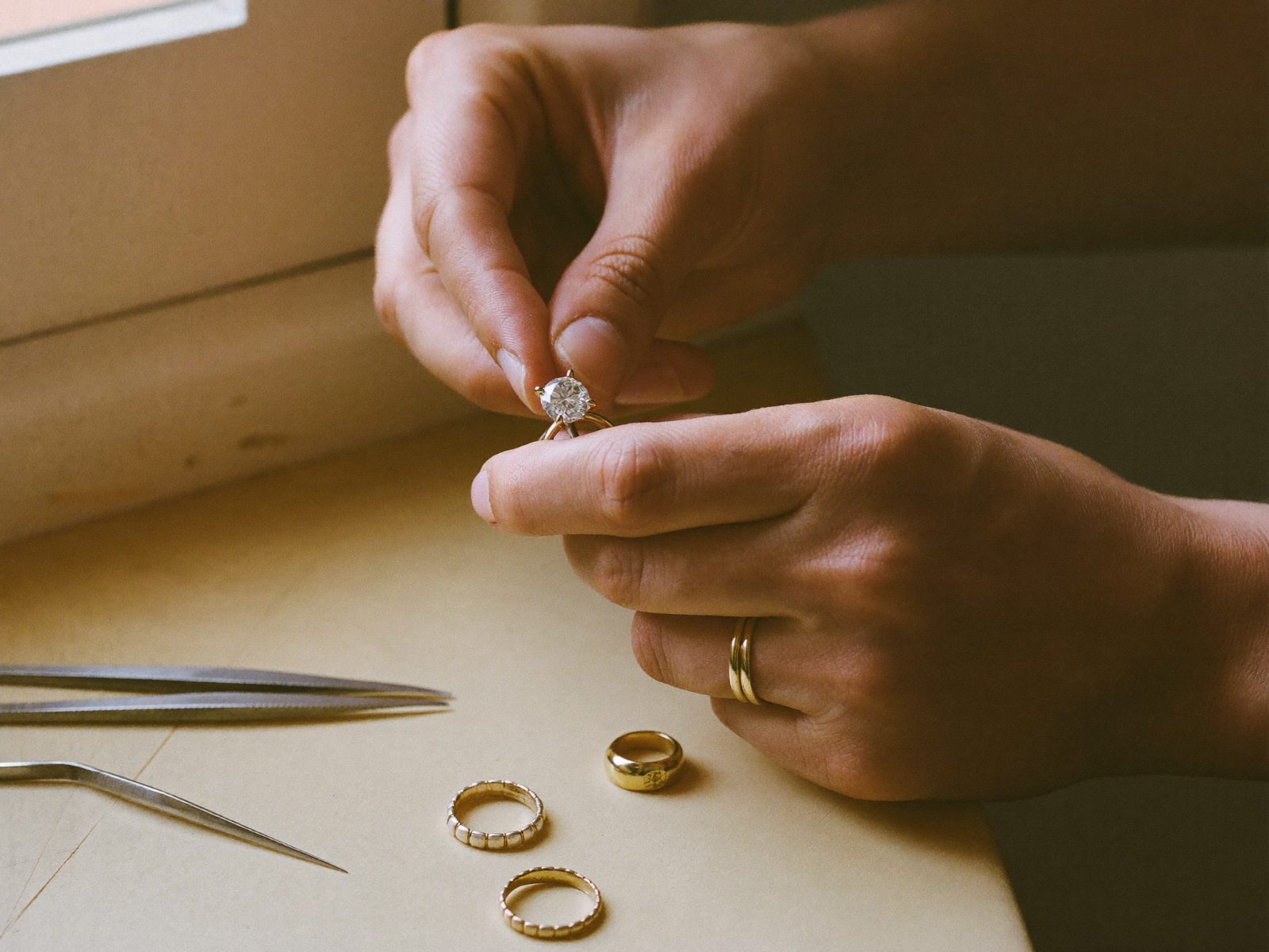 A jeweller's hands holding a round-brilliant solitaire ring over a workbench, with tweezers and three gold bands beside them