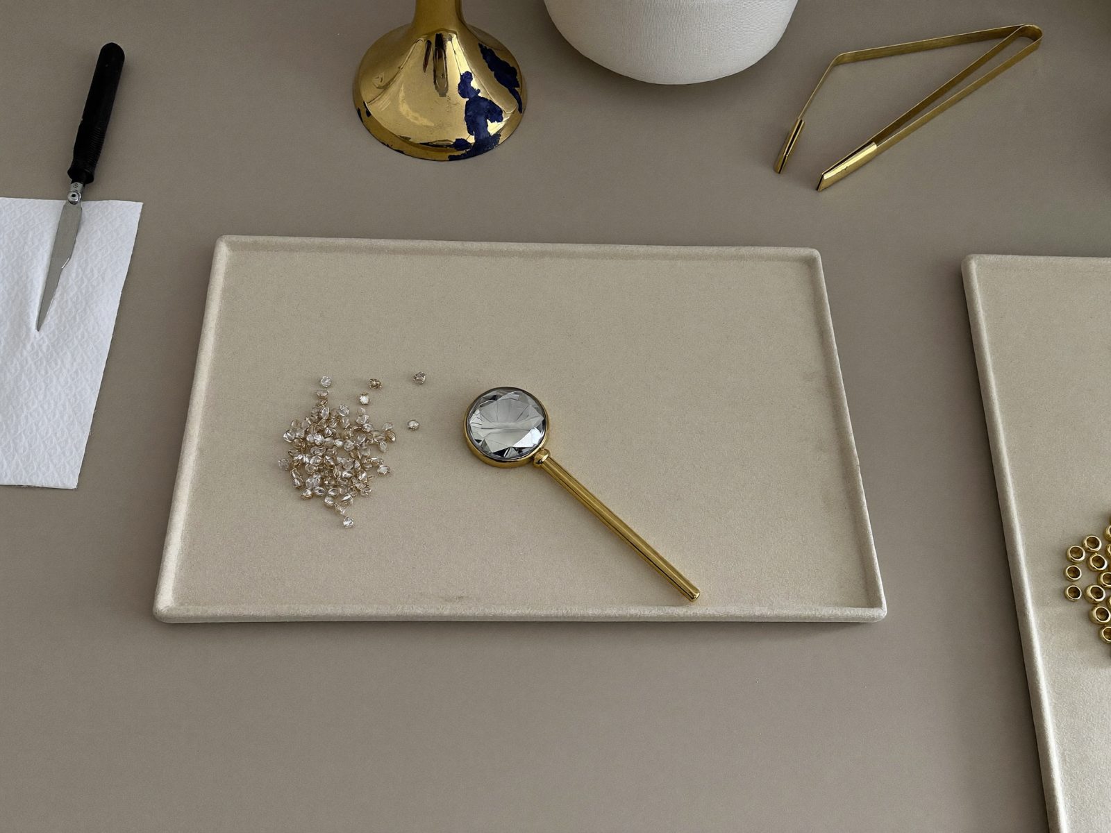 Loose lab-grown diamonds scattered on a cream suede tray beside a gold-handled loupe and brass tweezers