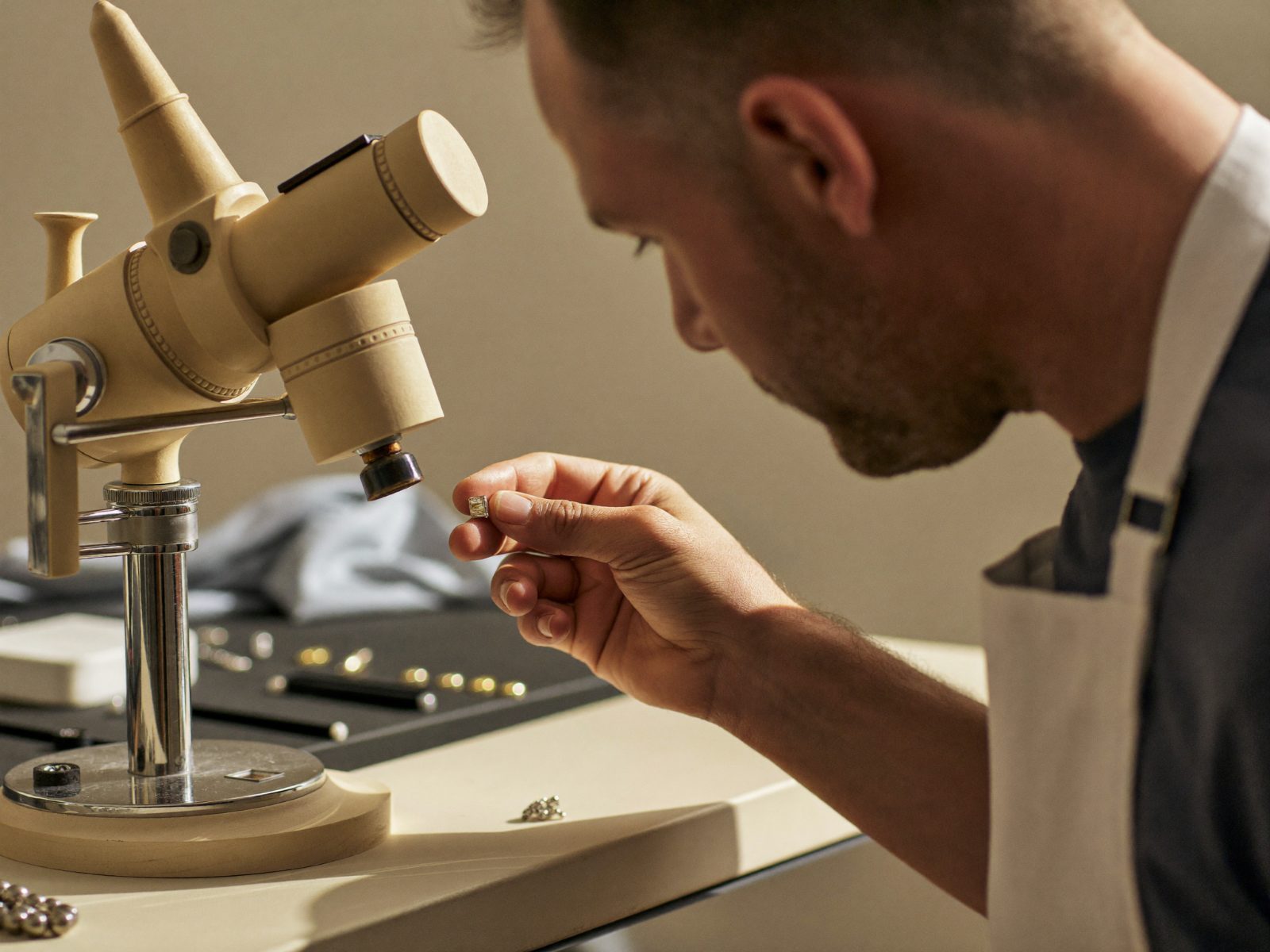 A jeweller in a linen apron examining a loose stone at a gem microscope, with a tray of stones behind
